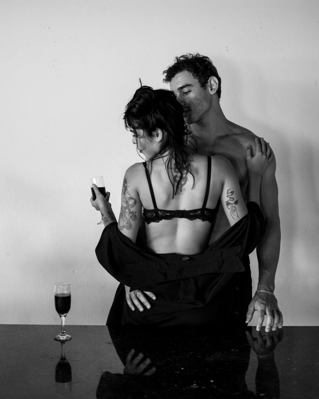 A tattooed couple embraces shirtless with wine in a moody black and white setting.