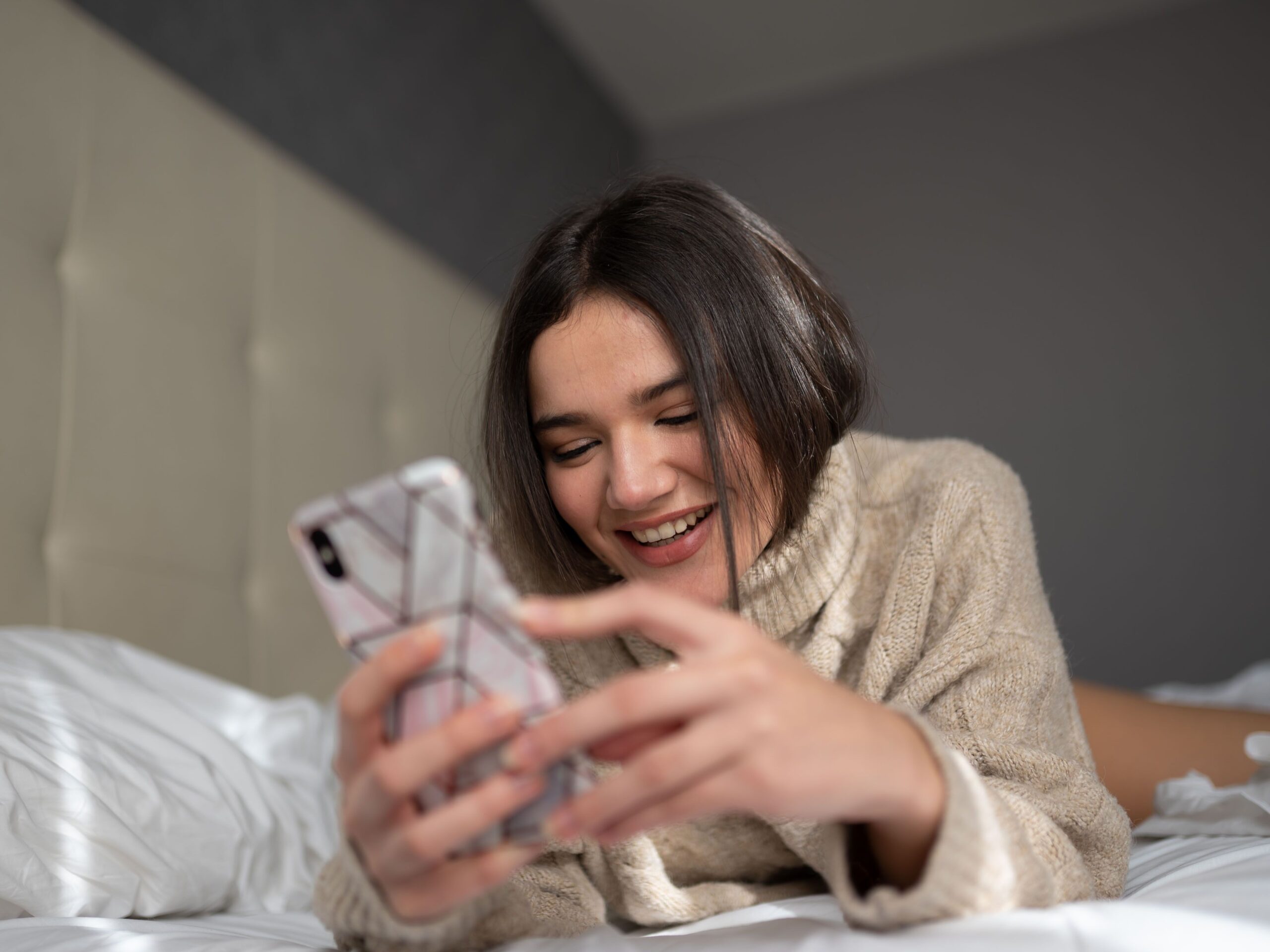 Low angle of positive young female millennial with dark hair in warm sweater lying on comfortable bed and smiling while messaging on mobile phone in sunny morning